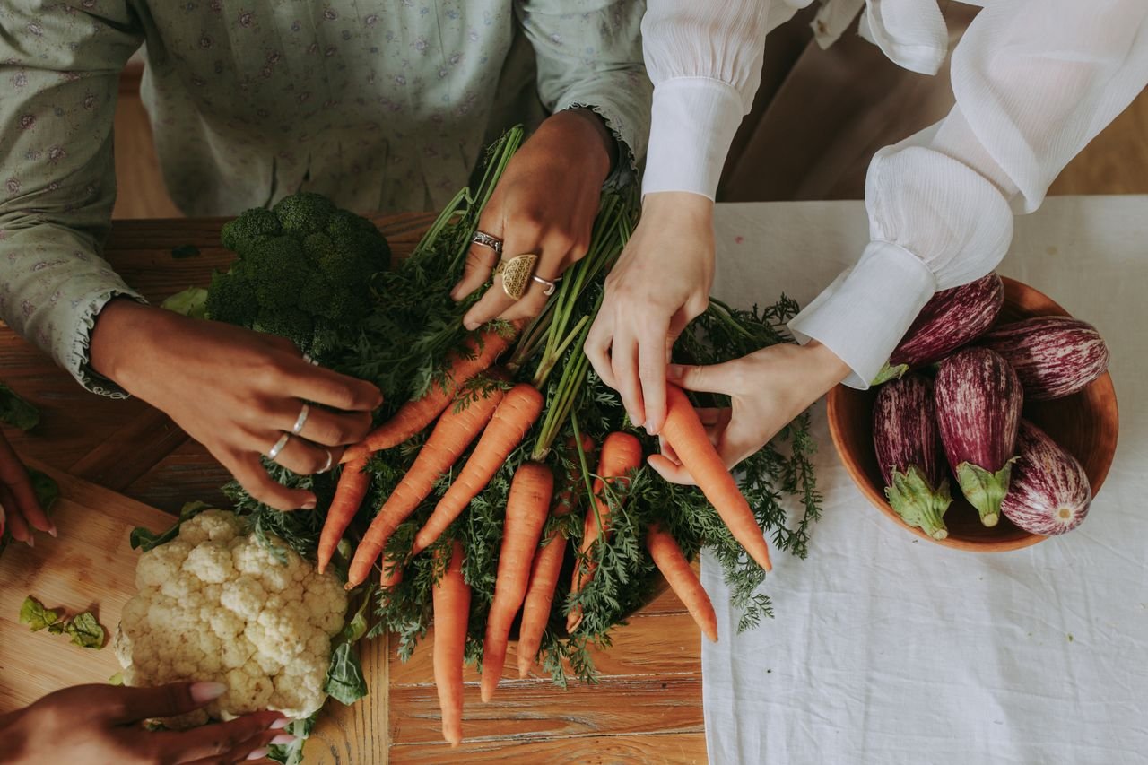 Diverse hands assorting fresh vegetables like carrots and eggplants on a rustic wooden table.